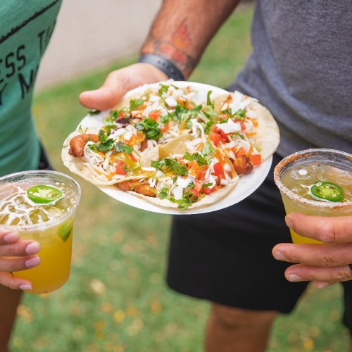 Two people hold drinks while sharing a plate of tacos topped with lettuce, tomatoes, and cheese, set on a grassy background.