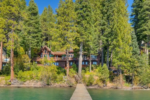 A wooden dock leads to a lakeside cabin surrounded by tall trees and clear blue water.
