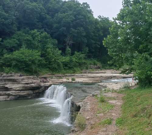 A serene waterfall cascades into a calm river, surrounded by lush green trees and rocky terrain.