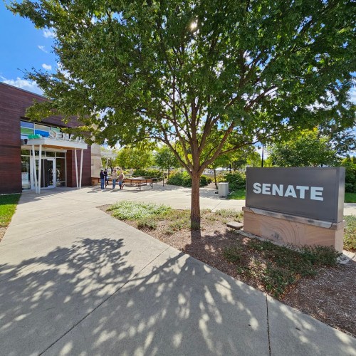 A modern building with a sign reading "SENATE," surrounded by trees and a clear blue sky. People are gathered outside.
