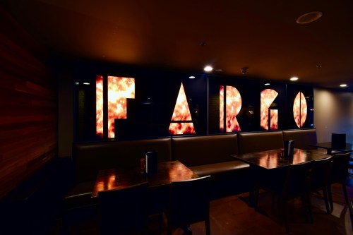 A dimly lit restaurant interior featuring the word "FARGO" in illuminated letters on a wall, with wooden tables in view.