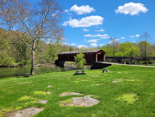 A scenic view of a red covered bridge over a river, surrounded by green grass and trees under a blue sky with clouds.