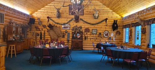 Interior of a rustic lodge with wooden walls, decorated with taxidermy, featuring tables set for dining.