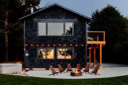 A modern black house with large windows, surrounded by trees, features a fire pit and seating area in the yard.