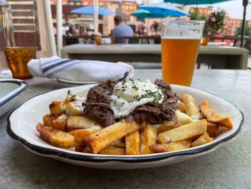 A plate of fries topped with shredded beef and a fried egg, with a glass of beer in the background.