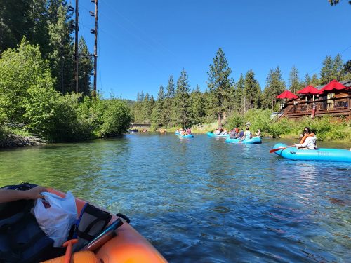 A group of people kayaking on a clear river surrounded by trees and a wooden structure in the background.