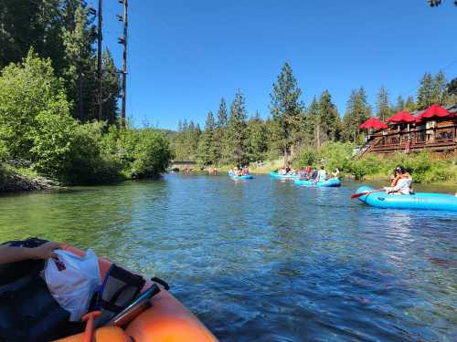 A group of people kayaking on a clear river surrounded by trees and a wooden structure in the background.