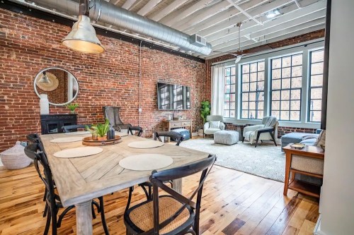 A stylish living space with exposed brick walls, a wooden dining table, and cozy seating areas by large windows.