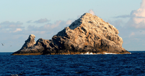 A rugged rock formation rises from the ocean, surrounded by water and a clear sky with scattered clouds.