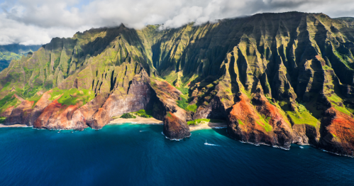 Aerial view of lush green mountains and rugged cliffs meeting a blue ocean under a partly cloudy sky.