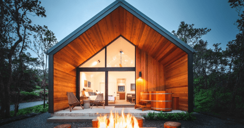 A modern wooden cabin with a fire pit in front, surrounded by trees, showcasing a cozy interior through large windows.
