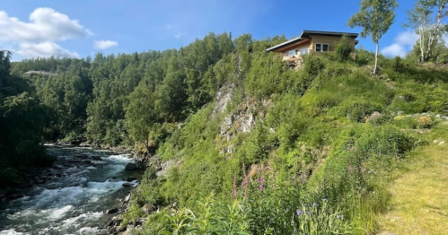 A house perched on a green hillside overlooking a flowing river, surrounded by lush trees and vegetation.