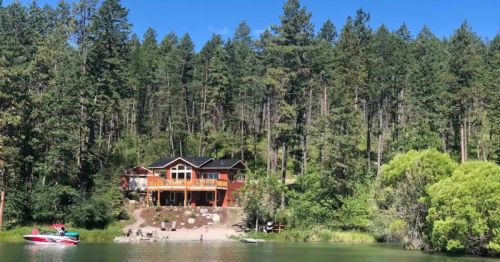 A lakeside cabin surrounded by tall trees, with a small boat and seating area by the water. Clear blue sky above.