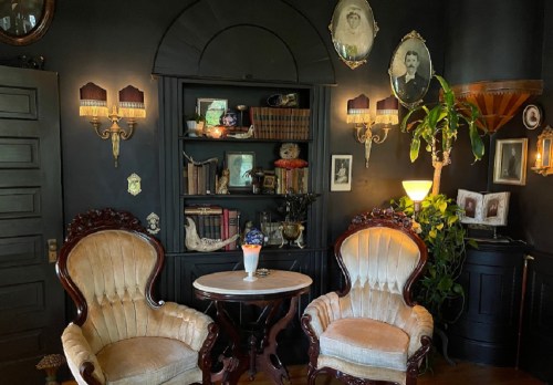 Cozy vintage sitting area with two plush chairs, a small table, and a dark bookshelf adorned with decor and framed photos.