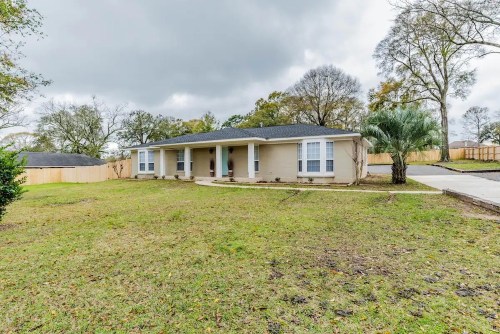 A single-story beige house with a front yard, surrounded by trees and a wooden fence, under a cloudy sky.