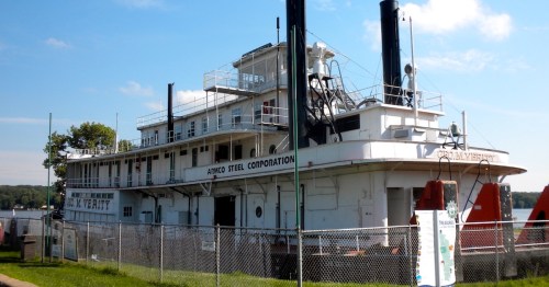 A historic steamboat docked by the river, featuring multiple decks and smokestacks, surrounded by a fence.