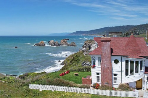 A coastal view featuring a white house with a red roof, green lawn, and rocky shoreline under a clear blue sky.