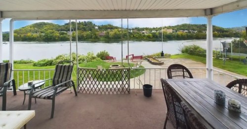 A covered patio with seating overlooks a river and green landscape, featuring a fire pit and distant hills.