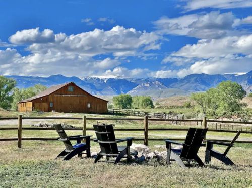 A scenic view of a barn with mountains in the background, featuring black chairs around a fire pit in the foreground.