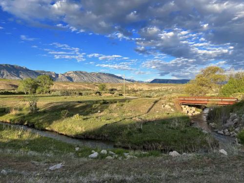 A serene landscape featuring a stream, grassy fields, and distant mountains under a partly cloudy sky.