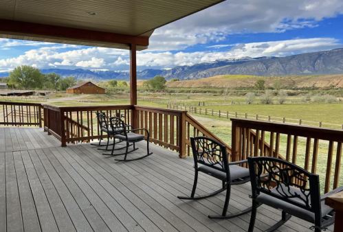 A scenic view from a porch with rocking chairs, overlooking a green field and mountains under a blue sky.