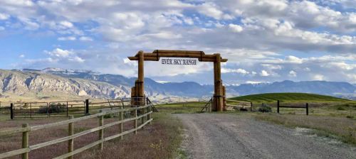 A wooden archway marks the entrance to a ranch, with mountains and a blue sky in the background.