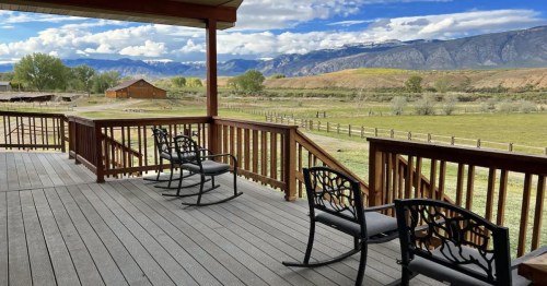 A scenic view from a wooden deck with rocking chairs, overlooking green fields and mountains under a blue sky.