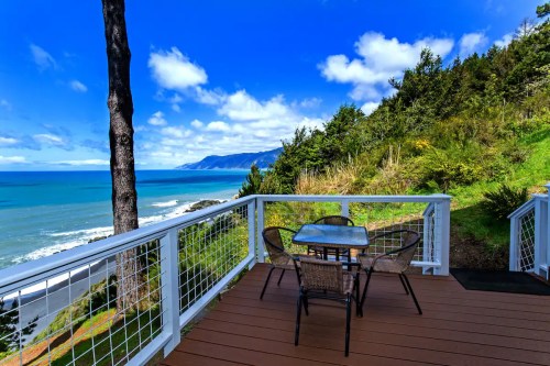 A scenic coastal view from a deck with a table and chairs, surrounded by greenery and blue skies.