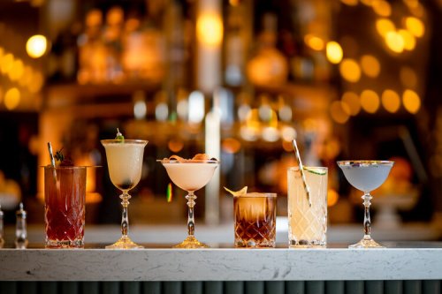 A row of six elegant cocktails on a marble bar, with a warm, blurred background of lights and bottles.