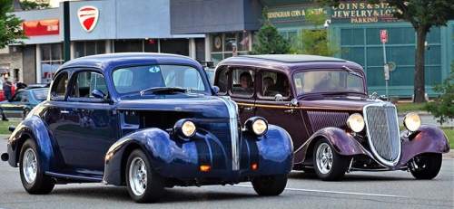 Two classic cars, one blue and one maroon, cruising down a city street with storefronts in the background.