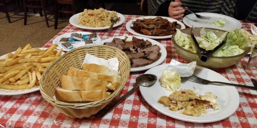 A table set with various dishes: rice, meats, salad, fries, bread, and utensils on a checkered tablecloth.