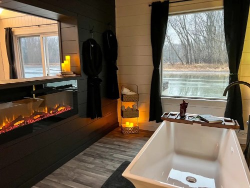 Cozy bathroom with a freestanding tub, fireplace, and large window overlooking a serene, wooded view.
