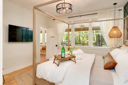 Cozy bedroom with a four-poster bed, TV, and large windows overlooking greenery, featuring a tray with drinks and flowers.