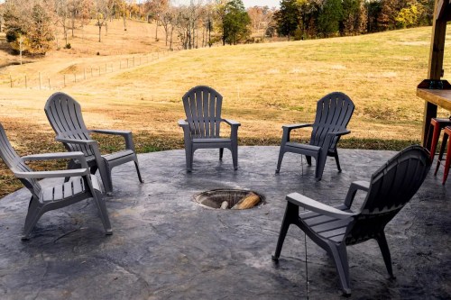 A circular patio with five gray chairs arranged around a fire pit, set in a grassy landscape with trees in the background.