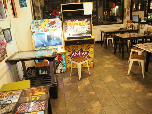 A cozy gaming cafe with arcade machines, tables, and colorful decor, featuring a "Marvel vs. Capcom" arcade cabinet.