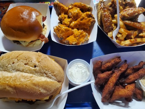 A variety of fast food: a burger, chicken wings with sauce, fries, and fried strips, all served in paper trays.