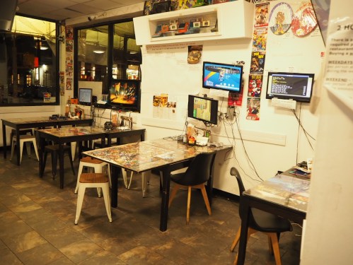A cozy gaming cafe with several tables, each equipped with monitors and gaming setups, surrounded by colorful decor.