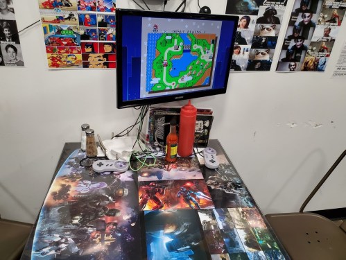 A gaming setup with a monitor displaying a Mario game, surrounded by posters and a condiment bottle on a themed table.