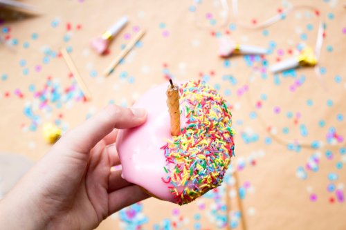 A hand holds a pink frosted donut with colorful sprinkles, surrounded by festive confetti and party decorations.