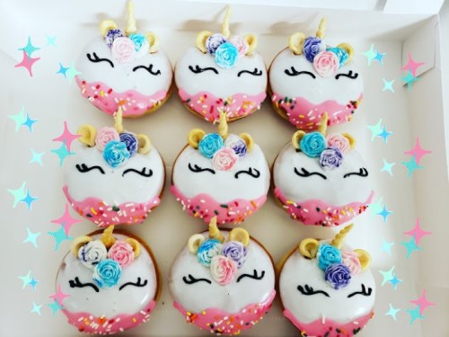 A box of colorful unicorn-themed donuts with pink icing, sprinkles, and decorative flowers on top.