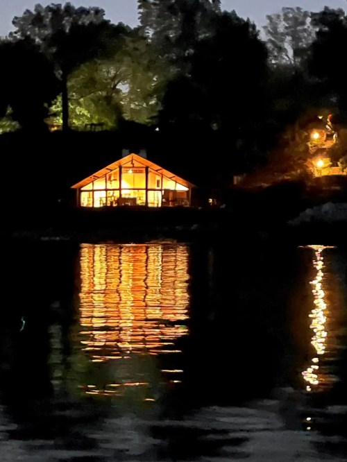 A lit cabin by the water at night, with reflections shimmering on the surface. Trees are silhouetted in the background.