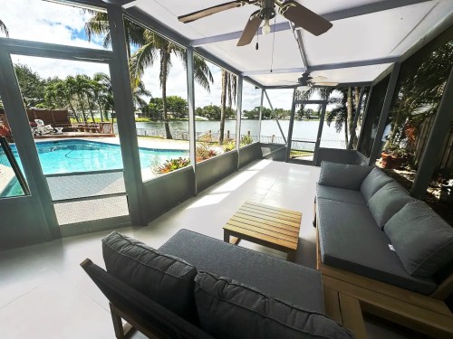 A screened patio with a sofa and coffee table, overlooking a pool and lake surrounded by palm trees.