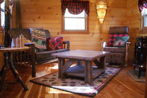 Cozy cabin interior with wooden walls, a rustic coffee table, two chairs, and decorative pillows. Warm and inviting atmosphere.