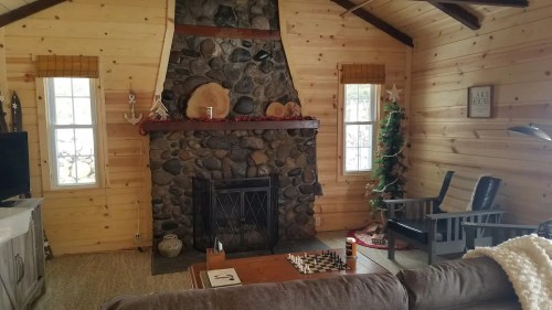 Cozy cabin interior with a stone fireplace, wooden walls, chess set on a table, and a decorated Christmas tree.