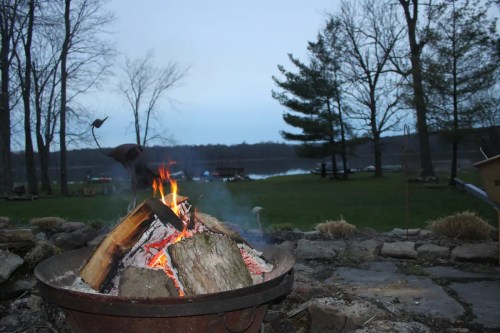 A cozy campfire burns in a fire pit, with trees and a lake visible in the background at dusk.