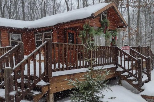 A cozy wooden cabin surrounded by snow, with a festive wreath and a small tree on the porch, amidst falling snowflakes.