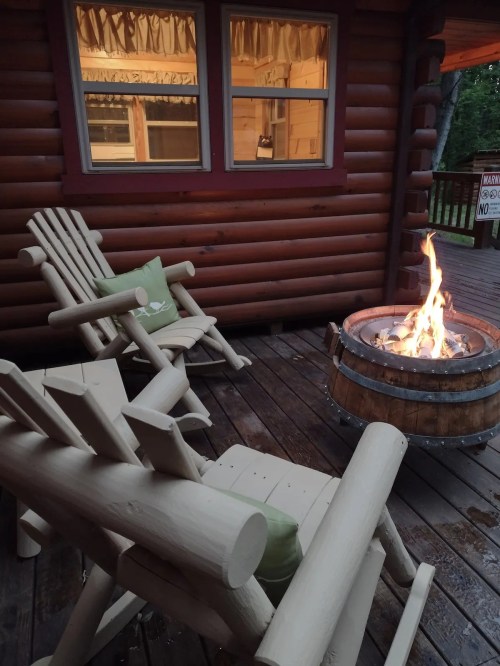 Two wooden chairs with green cushions beside a fire pit on a wooden deck, with a cabin window in the background.