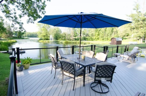 A serene deck with a blue umbrella, table, and chairs overlooking a pond surrounded by greenery.