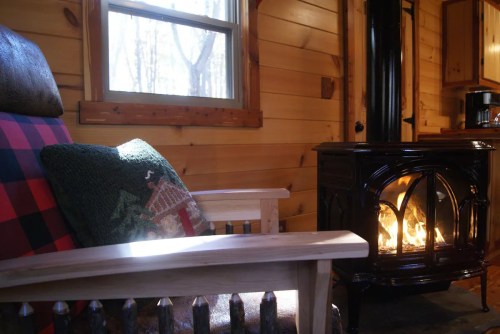 Cozy cabin interior with a wooden chair, decorative pillow, and a black stove emitting warm light.