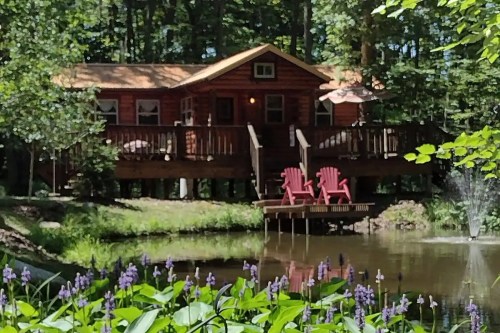 A cozy log cabin by a pond, surrounded by trees and flowers, with two red chairs on a deck.